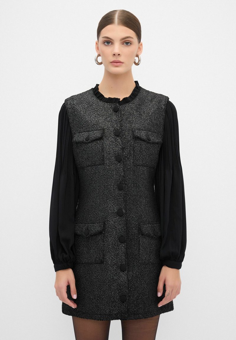 Vestido de tweed negro con escote fruncido, cuatro bolsillos y botones en la parte delantera. Mangas largas negras con textura plisada. Tejido suave y texturizado.