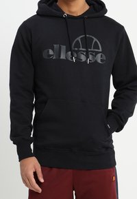 Osoba oblečená v černé mikině ellesse s přední kapsou na břiše a bordó kalhotách s malým logem na levém stehně.