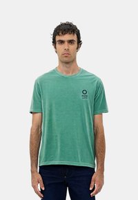 T-shirt in cotone verde con colletto rotondo, maniche corte e un piccolo logo circolare nero sul petto sinistro. Abbinato a jeans in denim scuri.