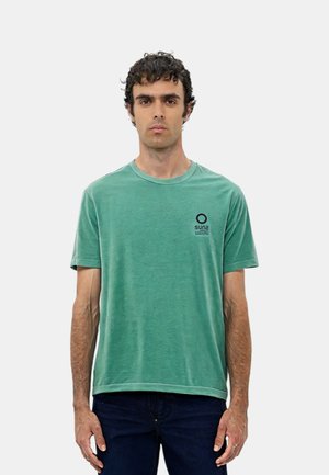 T-shirt in cotone verde con colletto rotondo, maniche corte e un piccolo logo circolare nero sul petto sinistro. Abbinato a jeans in denim scuri.