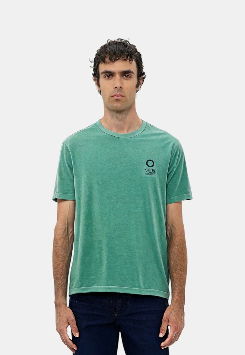 T-shirt in cotone verde con colletto rotondo, maniche corte e un piccolo logo circolare nero sul petto sinistro. Abbinato a jeans in denim scuri.