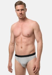 Mężczyzna bez koszulki, w jasnoszarych bokserek z czarnym paskiem, stojący na tle jednolitego, jasnoszarego tła.