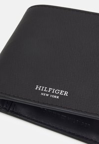 Portafoglio in pelle nera con superficie testurizzata. Caratterizzato da "HILFIGER NEW YORK" impresso in argento nella parte inferiore. Design pulito e minimale.