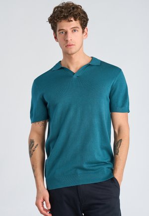 Giovane uomo con capelli ricci che indossa una polo a maniche corte lavorata a maglia di colore verde acqua e pantaloni scuri, in piedi con una mano in tasca.