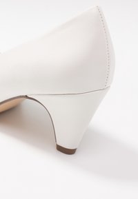 Weißer Leder-Pumps mit spitzer Spitze, strukturierter Oberfläche und einem zulaufenden Absatz mit braunem Boden. Gestochene Details an der Rückseite.