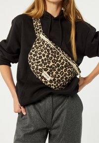 Sac en bandoulière à imprimé léopard en tissu beige et noir avec une fermeture éclair dorée, porté en travers du corps d'une personne sur un sweat à capuche noir.