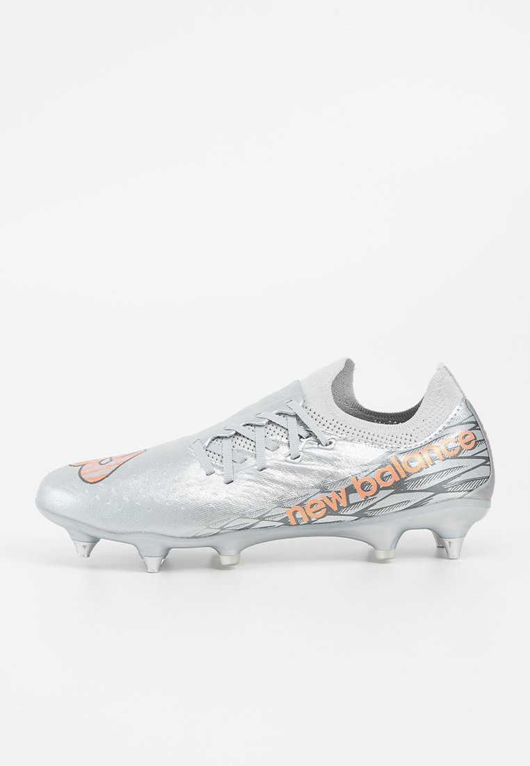 Crampons de football argentés avec une tige synthétique réfléchissante, accents noirs et logo "New Balance" orange, dotés de six crampons disposés de manière proéminente.
