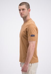 Rjava bombažna majica s kratkimi rokavi, okroglim ovratnikom in modro značko z logotipom na levi rokavi, prikazana na enobarvnem ozadju.