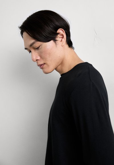 Schwarzer Strickpullover mit Rundhalsausschnitt, langen Ärmeln und strukturiertem Stoff. Model steht seitlich vor einem schlichten Hintergrund.