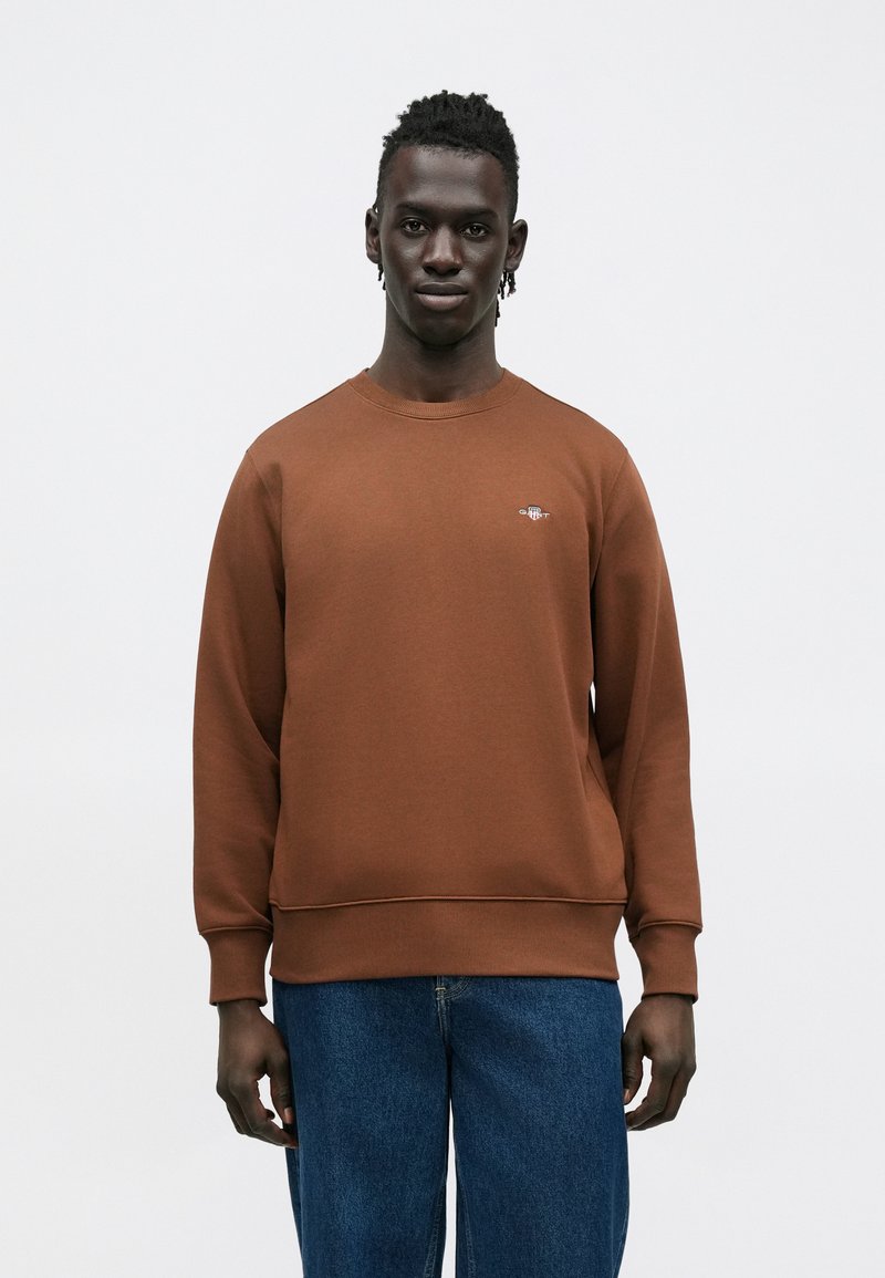 Sweat-shirt marron avec un col rond, des poignets et un ourlet côtelés. Présente un petit logo sur la poitrine. Associé à un jean bleu ample.