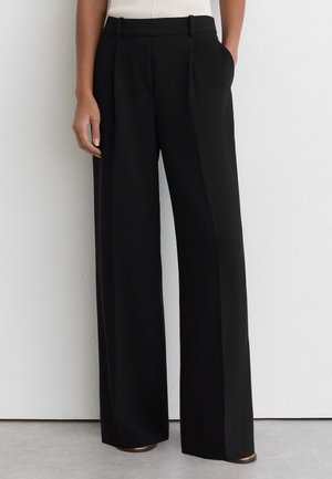 Pantalones negros de tiro alto y pernera ancha con pinzas delanteras, bolsillos laterales y trabillas para cinturón, combinados con una camiseta blanca metida dentro.