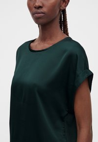 Primo piano di una persona con capelli intrecciati che indossa una maglietta in raso verde scuro a maniche corte con polsini risvoltati e scollo tondo su uno sfondo uniforme.