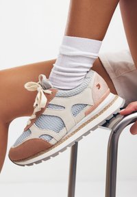 Sneaker leggera con tomaia in rete, accenti in suede beige e rosa, lacci crema e suola testurizzata. Presenta un dettaglio del logo sul lato.