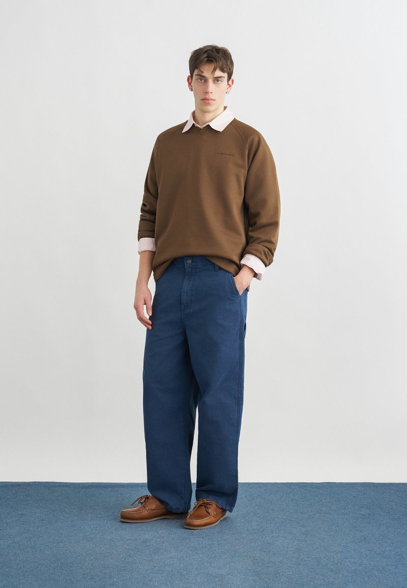 Jeune homme debout portant un sweat-shirt marron par-dessus une chemise rose, un pantalon bleu ample et des chaussures en cuir marron sur un sol recouvert de moquette bleue.