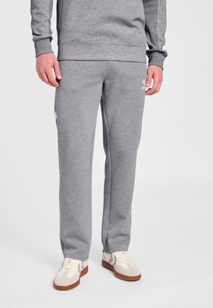 Mand iført lysegrå træningsbukser og matchende sweatshirt med hvide og beige sneakers, stående foran en ensfarvet baggrund.
