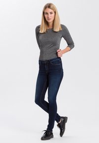 Grijs langmouwtop met subtiele glans, gedragen met aansluitende donkerblauwe jeans en zwarte veterschoenen. Achtergrond is plain wit.