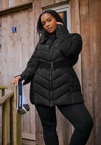 Schwarze Pufferjacke mit Zickzacksteppung, in der Taille mit einem Gürtel gesichert. Model hält eine Tasche mit "FINDWAY" auf dem Riemen.