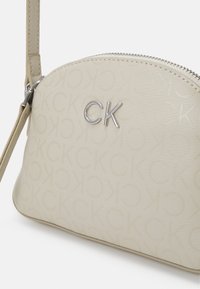 Béžová malá crossbody taška Calvin Klein se stříbrným logem CK, zapínáním na zip a nastavitelným popruhem, s embosovaným vzorem CK.
