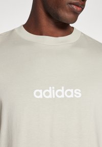 Světle béžové tričko vyrobené z bavlny s kulatým výstřihem. Na přední straně je výrazné bílé logo "adidas". Hladká textura.