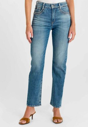 Vrouw draagt mid-rise blauwe rechte pijpen jeans gecombineerd met bruine open-toe sandalen met hak en een wit topje, staand tegen een effen achtergrond.