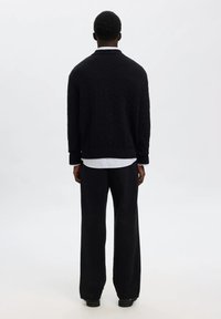 Suéter negro de punto con un cuello alto, usado sobre una camisa blanca a rayas; combinado con pantalones negros de corte holgado y zapatos negros.