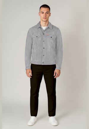 Mladý muž oblečený v světle šedé semišové bundě, černých kalhotách, bílých teniskách, stojící před jednoduchým světlým pozadím, čelem vpřed.