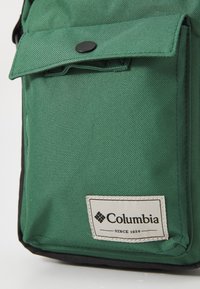 Borsa in tessuto verde Columbia con tasca frontale chiusa da un bottone a pressione nero e una patch bianca con la scritta "Columbia Dal 1938."
