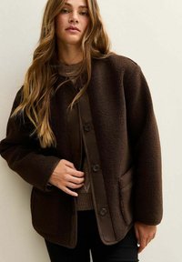 Chaqueta de forro polar marrón con una superficie texturizada, que cuenta con un cierre de botones, bolsillos laterales y ribetes marrones en contraste a lo largo de los bordes.