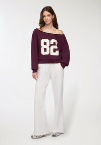 Felpa malva con spalle scoperte e stampa bianca "82", abbinata a pantaloni bianchi a gamba ampia e scarpe metallizzate, tessuto morbido, vestibilità rilassata.