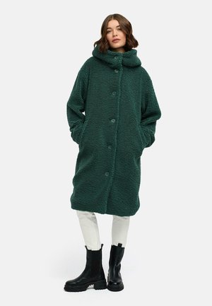 Manteau en polaire vert et moelleux avec une grande capuche pliée, des boutons sur le devant, des poches latérales et un design long et ample. Associé à un pantalon de couleur claire et des bottes.