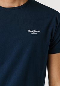 Tmavomodré tričko s krátkym rukávom, ktoré nosí osoba, s bielym logom "Pepe Jeans London" na ľavej strane hrude.