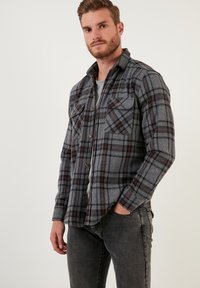 Camicia a quadri grigia con motivi rossi e neri, con bottoni e due tasche sul petto, realizzata in un tessuto testurizzato, indossata sopra una t-shirt grigia.