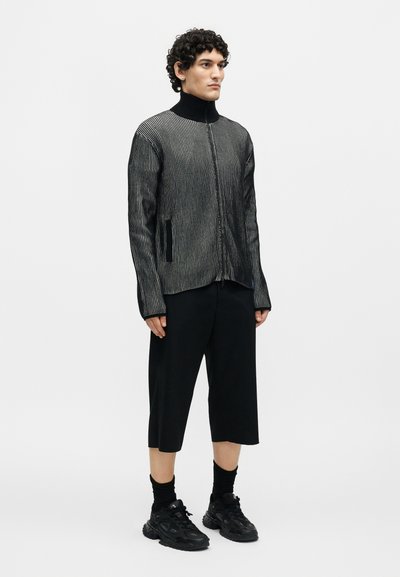 Άνδρας με σγουρά μαλλιά στέκεται φορώντας ένα μαύρο και γκρι ριγέ τζάκετ με φερμουάρ, μαύρο φαρδύ κοντό παντελόνι, μαύρες κάλτσες και μαύρα αθλητικά παπούτσια.