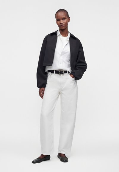 Personne portant une veste noire courte, une chemise blanche boutonnée, un pantalon blanc taille haute à jambes larges, une ceinture noire et des ballerines noires, debout avec une main dans la poche.