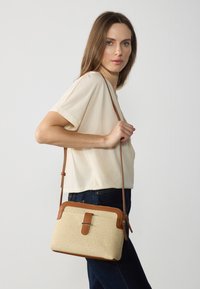 Donna che indossa un top corto color crema e jeans scuri tiene una borsa a tracolla intrecciata color sabbia e beige davanti a uno sfondo semplice.