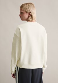 Kobieta z krótkimi blond włosami, ubrana w prostą, beżową bluzę i czarne spodnie z białymi paskami, stojąca tyłem na neutralnym tle.