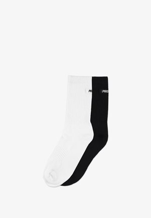 Skarpety z mieszanki bawełny w kolorze białym i czarnym, o prążkowanej fakturze, długości do połowy łydki, z nazwą marki "PROS" na czarnej skarpecie.