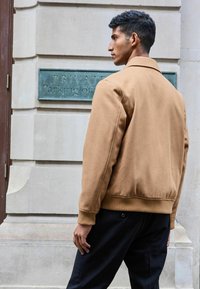 Chaqueta bomber de mezcla de lana marrón con puños y banda elástica acanalados, combinada con pantalones negros; posando frente a una pared de piedra texturizada.