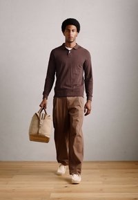 Uomo che cammina su un pavimento in legno indossando una camicia a maniche lunghe marrone, pantaloni khaki, scarpe bianche e portando una borsa beige nella mano destra.