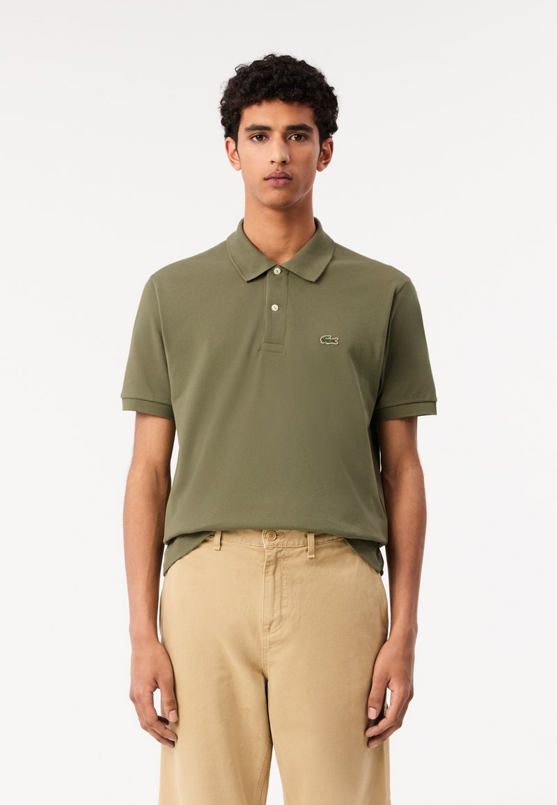 Olivgrön polo skjorta med klassisk krage och tvåknappsknäppning, prydd med en broderad logotyp på bröstet, i kombination med beiga byxor.