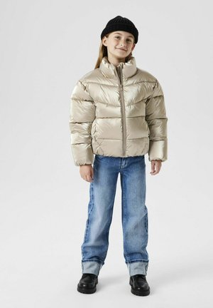 Giacca puffer beige con una texture lucida e trapuntata, collo alto e chiusura frontale con zip. Abbinata a jeans di jeans blu e scarpe nere.