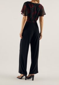 Combinaison noire avec manches transparentes à motif floral, offrant une coupe décontractée et une fermeture éclair au dos. L'ourlet inférieur est légèrement large, associé à des talons noirs.