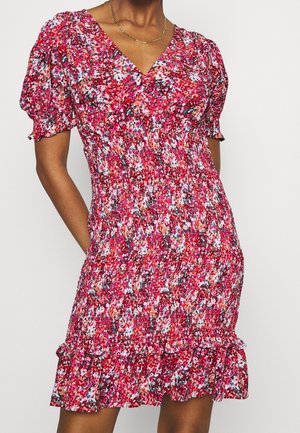 Žena v přiléhavých krátkých rukávech s květinovým vzorem v červené, růžové, bílé a černé barvě s volánkovým lemem, ruce v kapse.