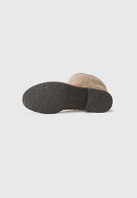 Zapato de ante marrón con una suela de goma negra texturizada que presenta patrones en forma de estrella. La puntera tiene forma redondeada y detalles cosidos.