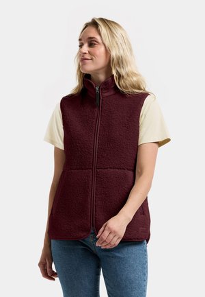 Blonde vrouw draagt een mouwloos bordeaux fleecevestje over een crème kortemouwen shirt en blauwe spijkerbroek, kijkt opzij.