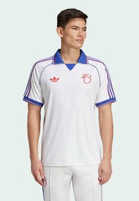 Biela športová košeľa s modrým golierom a červenými akcentami. Ikonické tri pruhy na rukávoch a jemný vzor na látke. Zobrazené logo Adidas.