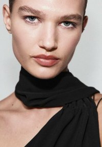 Retrato en primer plano de una persona con piel suave, que lleva una bufanda de tela negra caída sobre un hombro, destacando un color de labios suave y neutro y ojos azules.
