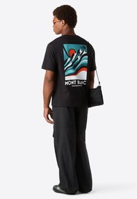 Hombre vistiendo una camiseta gráfica negra de Mont Blanc, pantalones negros holgados, gafas de sol naranjas y llevando una pequeña bolsa cruzada negra, mirando de lado.