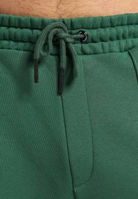 Des pantalons de survêtement verts en tissu texturé, avec une taille élastique et un cordon de serrage ajustable, ainsi que des poches latérales pour plus de fonctionnalité.