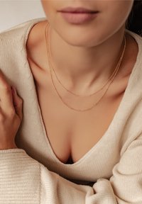 Goldene doppellagige Kette mit runden Perlen, getragen über einem beigefarbenen V-Ausschnitt-Pullover. Glatte Textur, zarte Kette und dezenter Glanz.